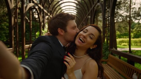Pov shot of sweet bride showing tongue on camera outdoors. Playful man and woman making facial expressions under arch. Portrait of cute couple doing selfie in park.