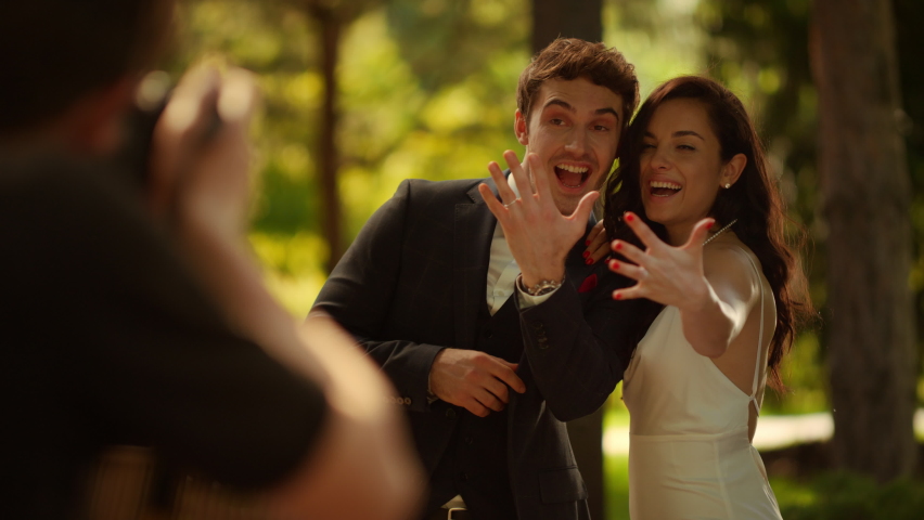 Happy couple posing for photograph on wedding photo session outdoors. Closeup joyful bride and groom showing rings on photo camera. Smiling man and woman smiling after wedding ceremony in garden.