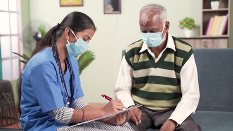 Young doctor or nurse writing prescription during home visiting to sick elder man while both worn face mask due to coronavirus covid-19 pandemic