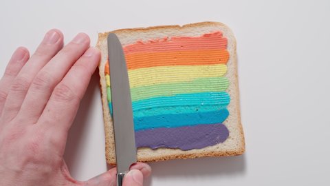 Rainbow. Male spreading colourful paste on a toast with knife. Top view.