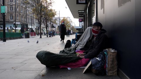 LONDON, ENGLAND, UNITED KINGDOM - CIRCA NOVEMBER, 2020: Two homeless people sitting on the ground by wall in UK capital city popular shopping street, Oxford street, during coronavirus pandemic.