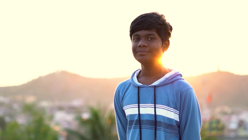 Portrait of an Indian kid smiling in front of the sunset. Happiness concept. Little kid smiles in front of sun depicting hope in the form of the light. Positive attitude of kid concept. | Shutterstock HD Video #1063704478