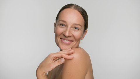 Close-up beauty portrait of young woman with smooth healthy skin, she gently touches her shoulders with her fingers on white background and smiles