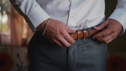 A caucasian man in gray pants and a white shirt and with a silver bracelet on his arm unbuckles and tightens the brown belt on his waist in the room