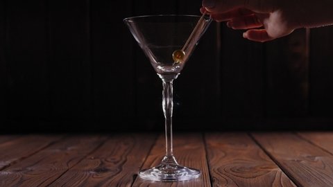 Close-up of a skewer with one olive falling into an empty Martini glass on a wooden surface on a black background. Slow motion.