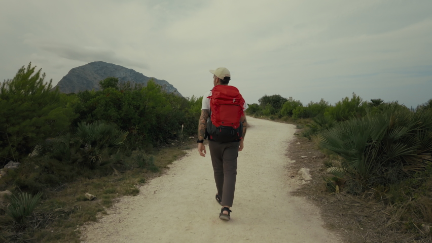 Adventure seeker, man with backpack walk on designated hiking path through national park. Young man, traveller turns around to make photo with camera. Hipster tourist on hike