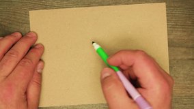 A man's hand writes with a green felt-tip pen on a cardboard sheet let it show. Work using a wooden Christmas table. Man makes manuscript congratulation Happy New Year. Flat laid writing items.