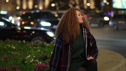 portrait of a beautiful young woman resting on a bench in the center of the city, in the background cars are passing by and the city lights are shining. one in a big city.
