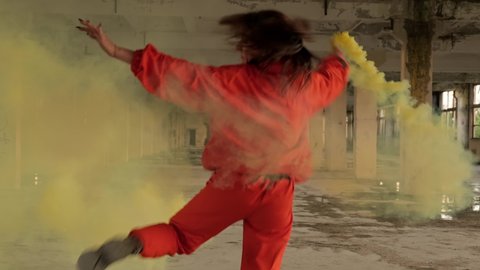 Stylish woman with smoke in hand dancing in abandoned factory. Freestyle dance improvisation. Young carefree woman in orange wear enjoys dancing in 4K, UHD