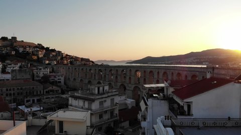 Aerial of Kavala aqueduct during summer sunset. The golden rays slowly penetrate through the buildings. A ship arrives at the sea port in the distance .