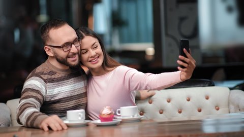 Happy couple posing taking selfie together use smartphone enjoying romantic date at cafeteria. Smiling man and woman photographing on mobile having coffee break. Shot on RED Raven 4k Cinema Camera