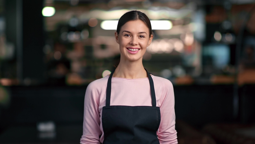 Portrait of friendly smiling woman in apron worker of cafe posing having positive emotion. Adorable female waiter or barista in uniform standing at cafeteria or restaurant. Medium shot on RED camera
