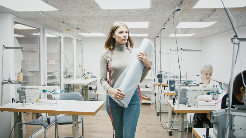 Sewing workshop. Young woman, tailor or fashion designer with tape measure is taking a roll of fabric and walking away past mixed race seamstresses working sitting at sewing machines. Slow motion Royalty-Free Stock Footage #1063722745