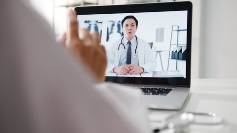 Young Asia male doctor in white medical uniform using laptop talking video conference call with senior doctor at desk in health clinic or hospital. Social distancing, quarantine for corona virus.