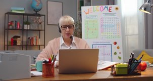 Smart Caucasian female teacher having class with students while staying at home. Confident adult woman in glasses and headphones teaching via video conference chat indoors. Technology, education.