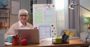 Smiling Caucasian woman wearing glasses and conferencing on laptop indoors. Adult female English teacher having online class and pointing to white board. Technology, education concept.