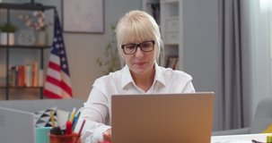 Confident adult female teacher working with laptop while sitting at table indoors. Thoughtful Caucasian woman typing on computer and putting off glasses. Technology, telework concept.