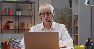 Smart adult woman in headphones working with laptop in homme office room. Caucasian female teacher in glasses conferencing with student and having online class. Education, e-learning concept.