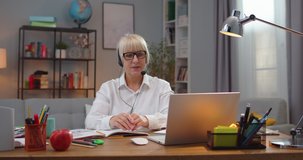 Professional female school teacher in glasses using laptop to video conference with students indoors. Adult Caucasian woman in headphones having online class. Pandemic, education, technology.