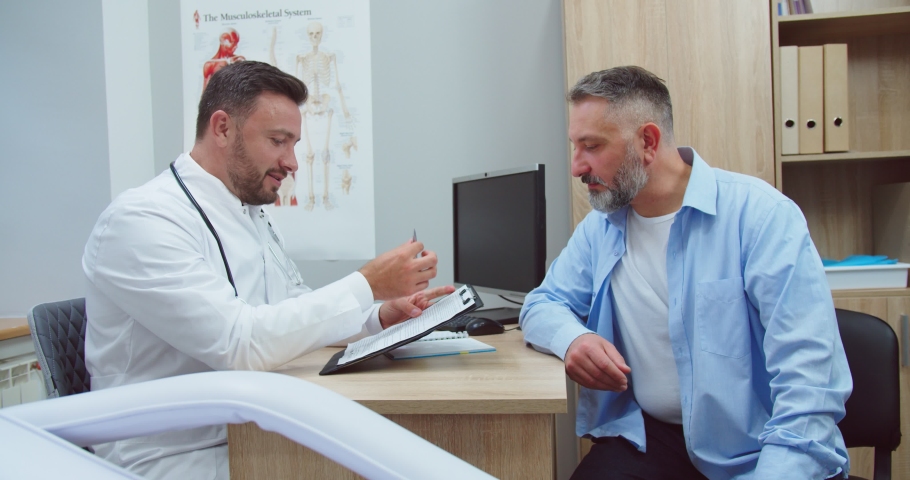 Portrait of Caucasian middle-aged male patient entering hospital cabinet and giving medical history to man doctor. Handsome physician giving advice to patient on consultation in clinic. Health concept | Shutterstock HD Video #1063727815