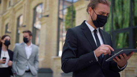 Confident businessman in mask working on tablet while going to work outdoors. Masked man using digital device at city street. Business people maintaining social distance with colleagues outside.