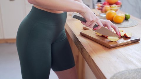 Close up Female hands cut fruit with a knife on a board table in the kitchen. Unrecognizable fitness girl in sportswear slicing apple. Female prepares fruit smoothies or fresh juice for blender juicer