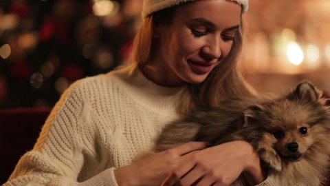 Cute Happy Young Woman is playing with her lovely little Dog, Puppy. Young Female is celebrating Christmas, New Year at Home with her Dog. Cozy Festive Evening. Happy Holidays. Christmas Spirit.