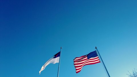 Two Flags are waving in the wind on side by side poles. One is a US Flag the other is the Ecumenical Christian Flag which has a cross on white background. That flag represents all denominations.