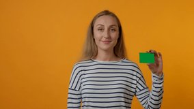 Portrait of a young smiling woman holding a blank green card in her hand. The concept of banking, credit card, business card holders. Yellow background. 4K video.