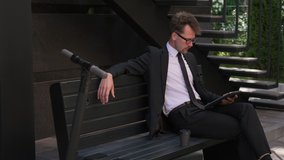 Businessman in suit sitting on bench next to paper cup of coffee in courtyard of office building, talking by online video call using tablet. Electric scooter nearby. Close up, slow motion