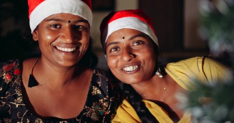 Close-up CC slow-motion handheld shot of two young Indian women smiling joyous good times wearing Santa hats decorating Christmas Tree, handling wrapped presents at dark night outdoors hugging share
