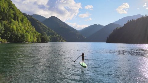 Female silhouette doing water sport during summer holidays. Concept of active tourism and travel. Aerial drone view of pretty asian Korean woman paddle on sup board at mountain lake during sunset.