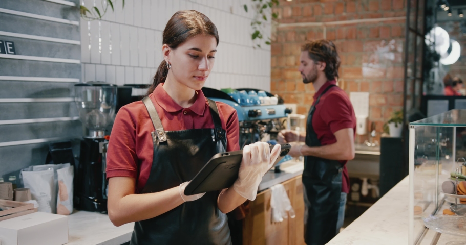 Young attractive female coffee store worker using digital tablet computer smiling to camera inside modern cafeteria. Occupation. Person portrait. Royalty-Free Stock Footage #1063746658