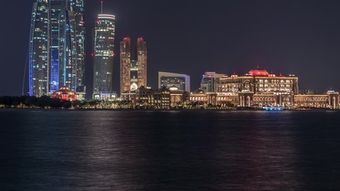 Skyscrapers of Abu Dhabi illuminated at night with towers buildings and emirates palace timelapse reflected in water. United Arab Emirates