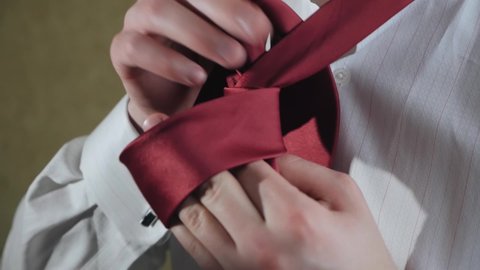 A man in a white shirt ties a red tie around his neck. learning tying a necktie