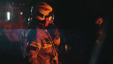 Close up of firefighter using radio to speak with his collegues. Fire engine with emergency lights and two more firemen in the background. High quality 4k footage
