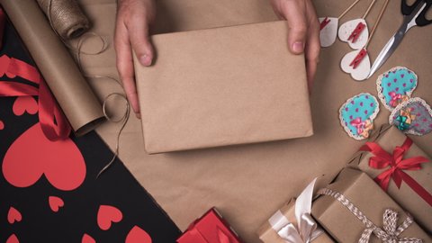 male hands put a box in craft paper on the table and start tying it with a string. The process of preparing gifts for the holiday. Valentine celebration concept