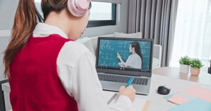 rear view of asian girl is learning math online through listening to female high school teacher teaching trigonometric function by laptop at home and write down the note