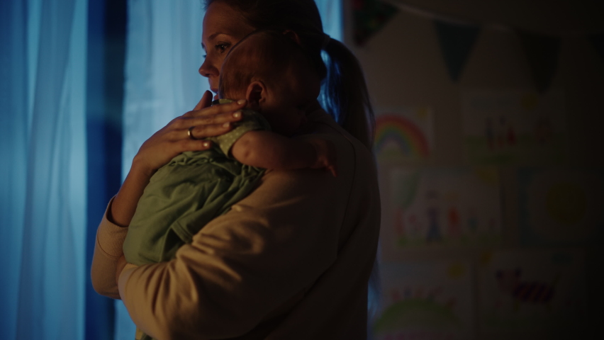 Happy Mother Holding Cute Newborn Baby in Cozy Dark Kids Bedroom in the Evening. Caring Mom Hugging the Neonate Toddler and They are Bonding Before Sleep. Concept of Childhood, New Life, Parenthood. Royalty-Free Stock Footage #1063762792