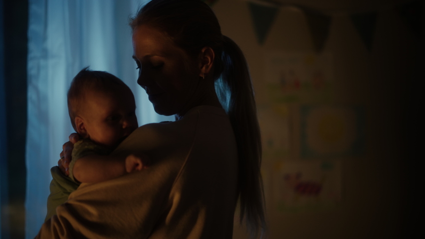 Happy Mother Holding an Adorable Newborn Baby in Cozy Dark Kids Bedroom in the Evening. Caring Mom and Neonate Toddler are Bonding at Home. Concept of Childhood, New Life, Parenthood. Royalty-Free Stock Footage #1063762807