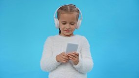 girl in fluffy sweater and headphones using smartphone isolated on blue