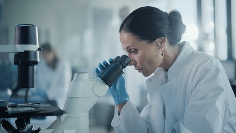 Medical Development Laboratory: Portrait of Beautiful Caucasian Female Scientist Looking Under Microscope, Analyzes Petri Dish Sample. Medicine, Biotechnology Research in Advanced Pharma Lab