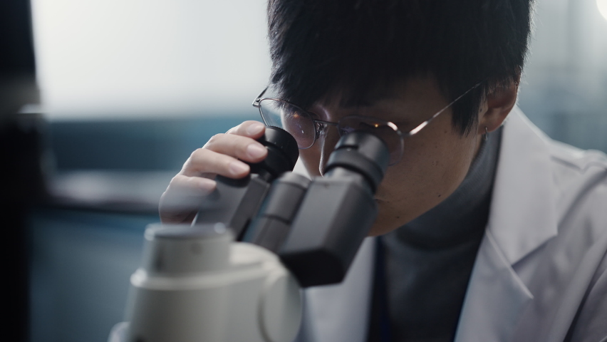 Medical Development Laboratory: Portrait of East Asian Scientist Looking Under Microscope, Analyzes Petri Dish Sample. Big Pharmaceutical Lab doing Medicine, Biotechnology, Microbiology,Drugs Research
