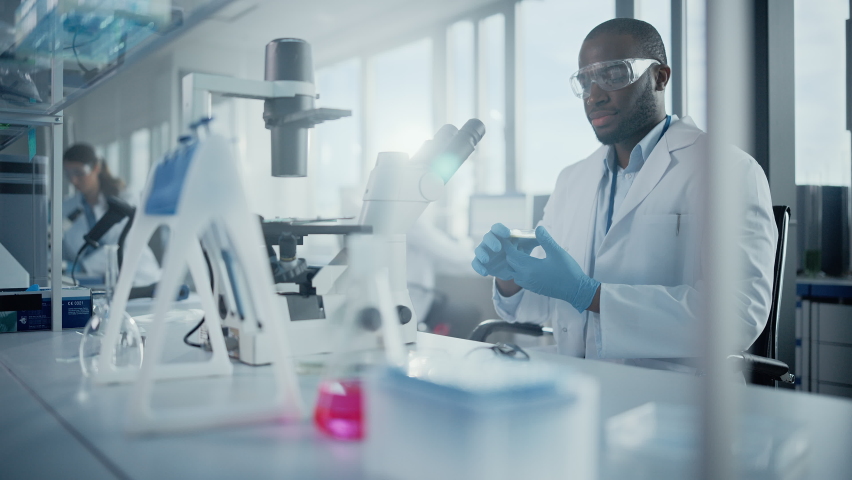 Medical Development Laboratory: Black Male Scientist Looking Under Microscope, Inspecting Petri Dish. Professionals Working in Advanced Scientific Lab doing Medicine, Vaccine, Biotechnology Research Royalty-Free Stock Footage #1063773790