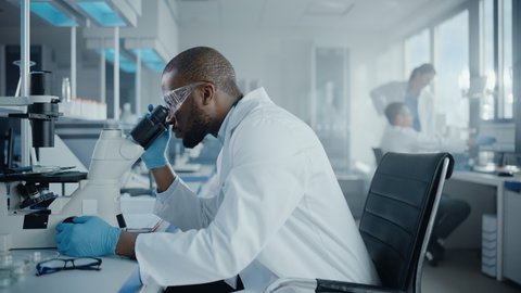 Medical Development Laboratory: Portrait of Black Male Scientist Looking Under Microscope, Analysing Petri Dish Sample. Professionals Doing Research in Advanced Scientific Lab. Side View Zoom In Shot