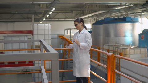 Technologist expert using tablet computer in chemical factory production line. Portrait of woman engineer in lab coat with digital tablet inspecting large industrial manufacture