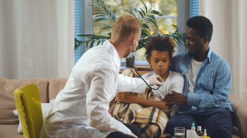 Doctor using stethoscope to listen heartbeat of child sitting with father in living room. Male pediatrician in protective mask examining afro schoolboy during home appointment