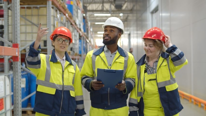 Warehouse workers team inspecting manufacturing storage. Multiethnic industrial colleagues in safety uniform and hardhat discussing distribution plan in large chemical warehouse | Shutterstock HD Video #1063780351