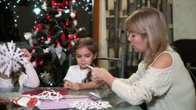 Mother and children sitting at table and cutting paper snowflakes, Christmas tree on background. Home video of happy family making handmade decorations together. Concept of holiday