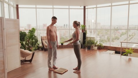 Full shot of young female yoga instructor explaining beginner basics of sadhu-yoga helping him to stand on nail board holding his hands